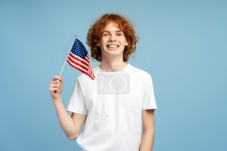 Téléchargez les photos : Joyeux garçon adolescent avec des cheveux bouclés rouges et des bretelles, tenant un drapeau des États-Unis, sur un fond de studio bleu. Concept d'immigration - en image libre de droit