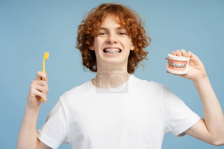 Happy, curly red haired male teenager wearing braces, holding a toothbrush and a model of jaws, against a blue background. Dental hygiene concept