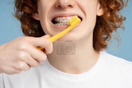 Close up shot of a ginger haired teen boy with braces, thoroughly cleaning his teeth, isolated on a blue background. Dental care practice concept