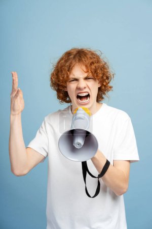 Téléchargez les photos : Photo verticale d'un adolescent furieux aux cheveux roux bouclés et aux accolades, criant à travers un haut-parleur haut-parleur, isolé sur un fond de studio bleu. Concept d'annonce - en image libre de droit