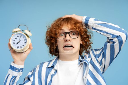 Photo for Portrait of humorous, curly haired redhead teen boy with braces, wearing glasses, showing alarm clock, and looking at camera in shock, isolated on blue studio background. Nerd late for school concept - Royalty Free Image