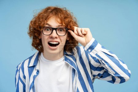 Closeup of a surprised ginger haired teenage boy with braces, wearing glasses and striped shirt, looking at camera, isolated on a blue studio background
