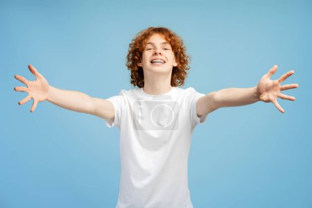 Foto de Sonriente y rizado adolescente pelirrojo con tirantes, brazos extendidos en una acogedora pose de abrazo, aislado sobre un fondo de estudio azul - Imagen libre de derechos