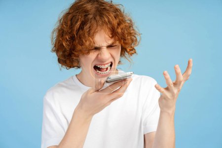 Photo for Snapshot of an angry teen with curly red hair and braces, shouting while holding his smartphone, engaged in an intense online conversation, isolated on a blue studio background - Royalty Free Image
