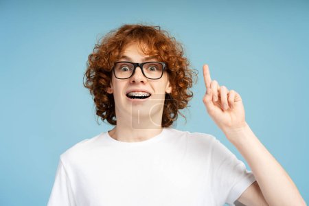 Téléchargez les photos : Portrait d'un adolescent roux aux cheveux bouclés et intelligent avec des bretelles, portant des lunettes et faisant le geste d'une idée avec son doigt, isolé sur un fond bleu. Essence d'une étude nerd concept - en image libre de droit