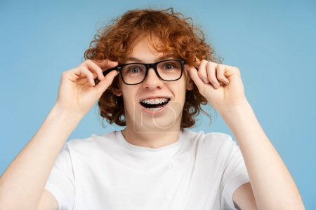 Foto de Imagen de un adolescente nerd de pelo rojo rizado con tirantes, equipado con anteojos, mirando directamente a la cámara con la boca ancha, aislado sobre un fondo azul - Imagen libre de derechos