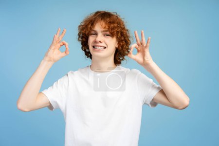 Téléchargez les photos : Adolescent heureux et confiant aux cheveux roux bouclés et aux bretelles, montrant le signe ok, isolé sur un fond bleu - en image libre de droit