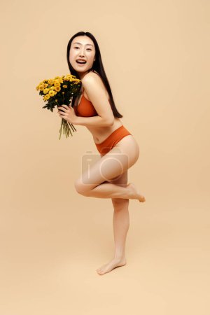Téléchargez les photos : Portrait heureuse séduisante femme asiatique confiante portant une lingerie sexy tenant de belles fleurs printanières isolées sur fond beige. Modèle de mode posant pour des photos. Journée internationale de la femme - en image libre de droit