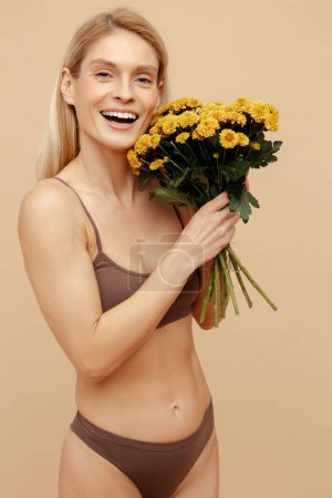 Téléchargez les photos : Portrait souriant belle femme tenant bouquet fleurs de printemps isolé sur fond beige. Joyeux mannequin de mode confiant portant une lingerie sexy posant en studio. Concept de journée internationale de la femme - en image libre de droit