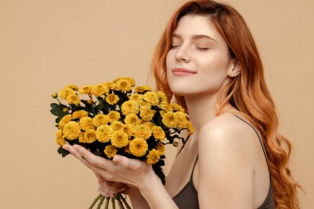 Foto de Retrato linda mujer de pelo rojo hermosa con los ojos cerrados sosteniendo ramo de flores de primavera aislados sobre fondo beige. Belleza natural, Día Internacional de la Mujer - Imagen libre de derechos