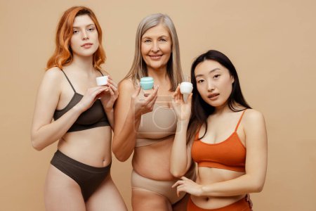 Téléchargez les photos : Groupe attrayant souriant femmes multiraciales portant des sous-vêtements sexy tenant des pots avec crème pour le corps isolé sur fond beige. Procédure beauté, concept de soins de la peau - en image libre de droit