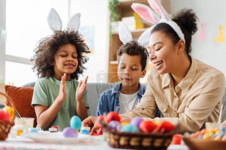 Photo for Smiling mother and cute kids wearing bunny ears painting easter eggs at home. Happy African American family celebration holidays. Sunday Easter concept - Royalty Free Image