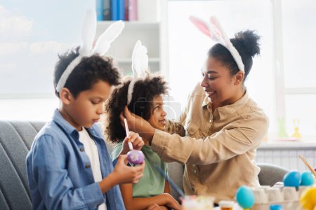 Photo for Smiling mother and cute kids wearing bunny ears painting and decorating easter eggs at home. Happy African American family celebration Easter together - Royalty Free Image