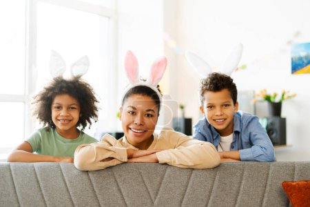 Téléchargez les photos : Portrait mère souriante avec des enfants portant des oreilles de lapin mignon regardant la caméra. Heureuse famille afro-américaine célébrer Pâques à la maison - en image libre de droit