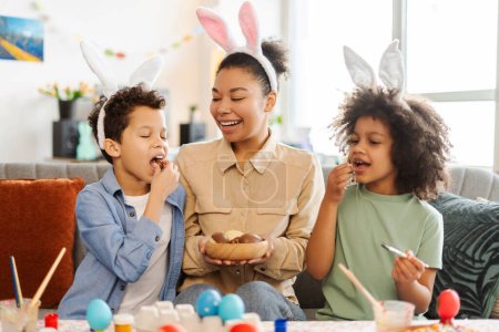 Foto de Retrato feliz familia latina comiendo huevos de chocolate en casa. Madre sonriente y niños con orejas de conejo celebran la Pascua juntos. Concepto de actividad vacacional - Imagen libre de derechos