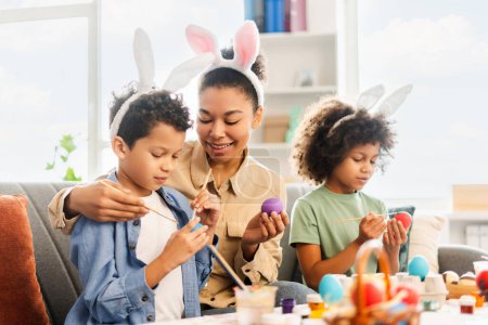 Photo for Smiling mother and children wearing bunny ears painting eggs at home. Happy African American family celebration Easter - Royalty Free Image