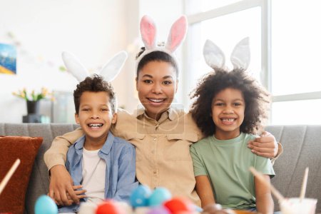 Foto de Madre sonriente retrato con niños con orejas de conejo mirando a la cámara. Feliz abrazo de la familia afroamericana celebrar la Pascua en casa - Imagen libre de derechos