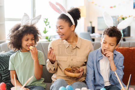 Photo for Portrait smiling African American family eating chocolate eggs at home. Happy mother and cute kids wearing bunny ears celebration Easter together - Royalty Free Image