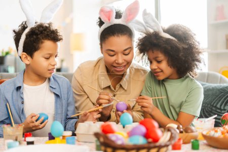 Foto de Linda familia latina sonriente: madre e hijos con orejas de conejo pintando huevos de Pascua en casa - Imagen libre de derechos
