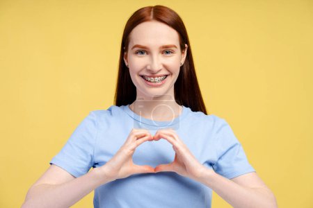 Happy ginger woman with braces showing heart shape for subscribers isolated on yellow background. Positive lifestyle, social media concept