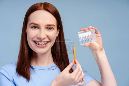 Foto de Retrato de una hermosa mujer pelirroja con tirantes sosteniendo cepillo de dientes, mirando a la cámara aislada sobre fondo de color azul. Concepto de rutina diaria e higiene matutina - Imagen libre de derechos