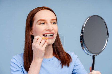 Foto de Atractiva mujer de jengibre mirando en el espejo, cepillándose los dientes con cepillo de dientes interdental aislado sobre fondo azul. Concepto de cuidado dental y salud - Imagen libre de derechos