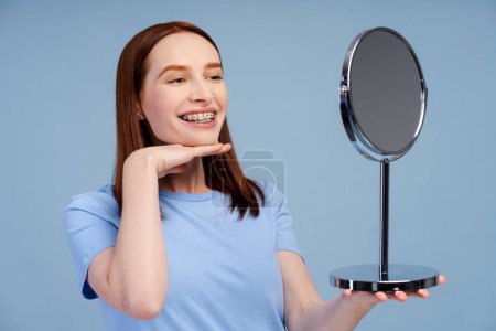 Photo for Attractive ginger woman looking in mirror, feeling satisfied with her teeth isolated on blue background. Concept of dental care and health - Royalty Free Image