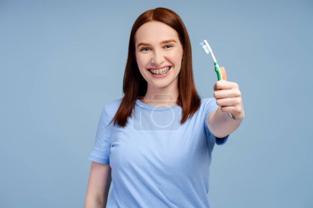 Porträt einer schönen Ingwerfrau mit Zahnspange, die eine Zahnbürste hält und die Kamera isoliert auf blauem Hintergrund betrachtet. Tagesablauf und morgendliches Hygienekonzept