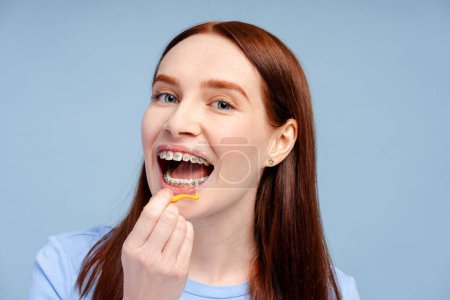 Foto de Primer plano de la mujer de jengibre con frenos que sostienen el cepillo de dientes, cepillándose los dientes en el baño en un fondo azul aislado. Concepto de cuidado dental - Imagen libre de derechos