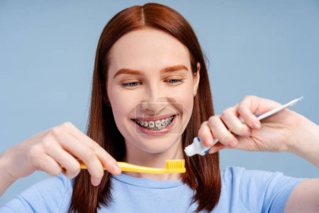 Photo for Beautiful red haired woman with braces holding pouring toothpaste on toothbrush isolated on blue colour background. Daily routine and morning hygiene concept - Royalty Free Image
