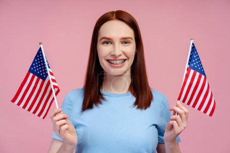 Téléchargez les photos : Portrait de femme excitée aux cheveux rouges tenant le drapeau américain, se réjouissant de la victoire, regardant la caméra isolée sur fond rose. Concept de jour de l'indépendance - en image libre de droit