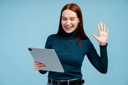 Téléchargez les photos : Belle femme aux cheveux rouges avec des accolades portant des lunettes tenant une tablette numérique, agitant la main, travaillant en ligne, ayant un appel vidéo. Technologie, concept de vidéoconférence - en image libre de droit
