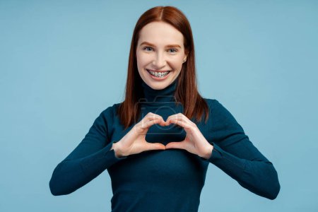 Happy ginger woman with braces showing heart shape for subscribers isolated on blue background. Positive lifestyle, social media concept