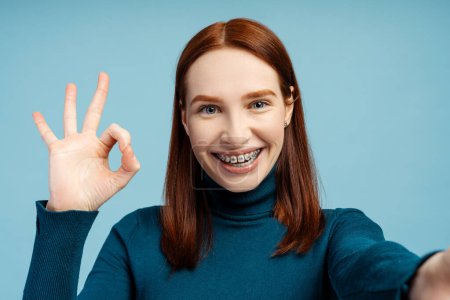Foto de Mujer jengibre sonriente con frenos dentales que muestran un signo aceptable mirando a la cámara haciendo selfie aislado sobre fondo azul. Emociones positivas, cuidado de la salud, concepto de ortodoncia - Imagen libre de derechos