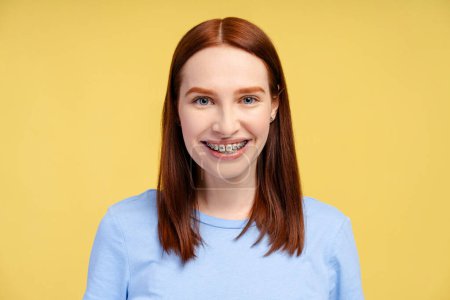 Téléchargez les photos : Photo d'une femme rousse souriante aux bretelles orthodontiques, portant un t-shirt bleu, regardant directement la caméra, isolée sur un fond jaune - en image libre de droit