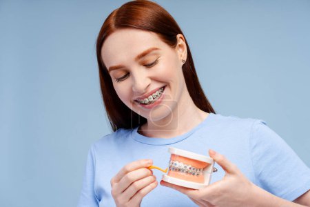Image of a smiling, attractive redhead woman demonstrating a tooth model cleaning with a floss holder, isolated on a blue background. Focuses on oral care
