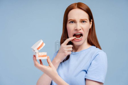 Portrait of a thoughtful redhead with braces, holding a dental structure model and lightly touching her teeth, isolated on a blue background. Dental hygiene concept