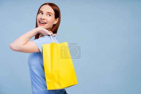 Smiling ginger haired lovely woman with dental brackets, holding a purchase-filled paper bag, isolated on a blue scene. Celebrating shopping spree concept