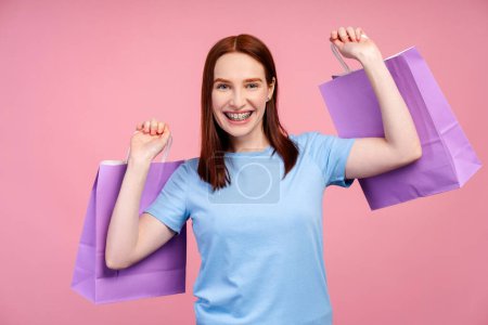 Foto de Mujer atractiva pelirroja sonriente con frenillos, agarrando bolsas de compras llenas de compras, aisladas sobre un fondo rosa. Concepto de compras - Imagen libre de derechos