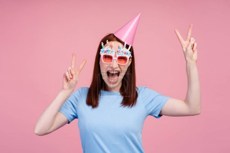 Téléchargez les photos : Image d'une fille aux cheveux roux joyeuse avec des appareils orthodontiques, célébrant son anniversaire avec un chapeau de fête et des lunettes amusantes, faisant des symboles de paix avec ses mains, isolée sur fond rose. Concept de parti - en image libre de droit