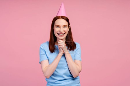 Schnappschuss einer begeisterten Frau mit kieferorthopädischen Brackets, die ihren Geburtstag mit einem Partyhut feiert, isoliert auf rosa Hintergrund. Festliche Idee