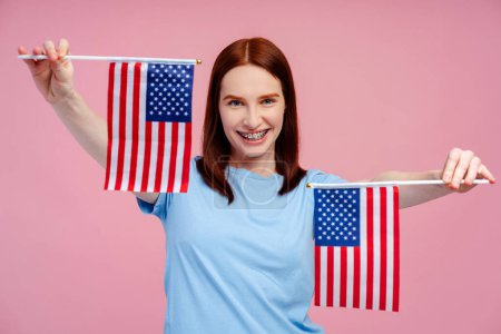 Attraktive Ingwerhaarige, die Klammern trägt und ein Paar amerikanische Flaggen in der Hand hält, isoliert auf rosa Hintergrund. Abstimmungskonzept