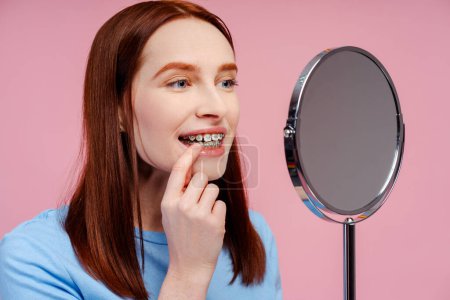 Foto de Imagen de una atractiva pelirroja con aparatos ortopédicos, examinando de cerca sus dientes en el espejo, aislada sobre un fondo rosa. Concepto de salud dental - Imagen libre de derechos