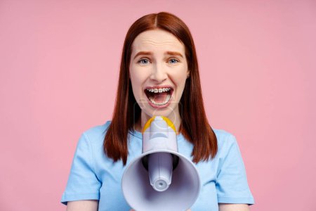 Photo for Closeup image of a beautiful ginger haired woman with braces, screaming through a megaphone, directly looking into the camera, isolated on a pink background. Announcement concept - Royalty Free Image