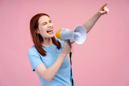 Red haired woman wearing brackets, energetically shouting through a loudspeaker, making a pointing gesture, isolated on a pink background. Promotion concept