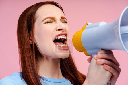 Closeup photo of a beautiful woman with ginger hair and orthodontic braces, vocally expressing through a megaphone, isolated against pink background. Announcement concept