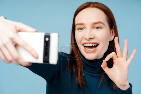Cheerful red haired woman with braces, wearing a cozy polo neck, taking a selfie on her mobile phone while making the OK gesture, isolated on blue background. Modern technologies concept