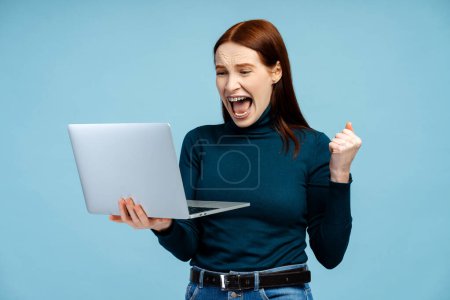 Téléchargez les photos : Photo d'une femme aux cheveux roux ravie avec des bretelles, tenant sur un ordinateur portable et faisant passionnément un geste de triomphe, isolée sur un fond bleu - en image libre de droit