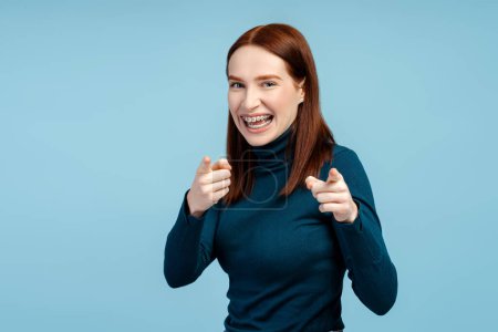 Photo for Cheerful red haired female with braces, in a sweater, making an optimistic finger gesture, eyes on the camera, isolated on a blue background - Royalty Free Image