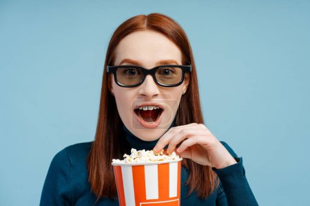 Photo for Portrait of an astonished ginger woman with braces, donning a polo neck sweater and 3D glasses, enjoying popcorn, mouth wide open, isolated on blue background - Royalty Free Image